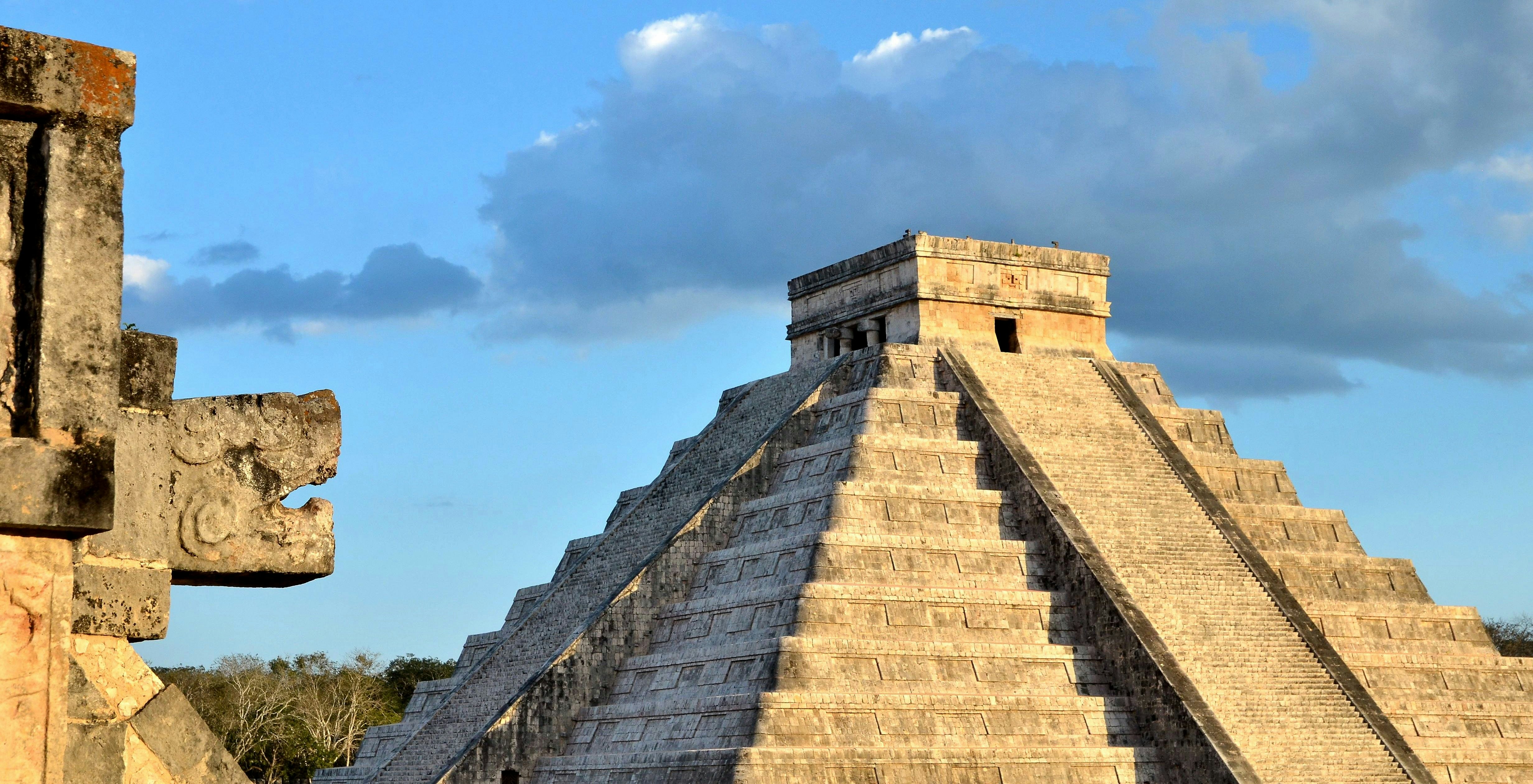 A large stone snake head faces towards the large stone temple of El Castillo in Mexico 